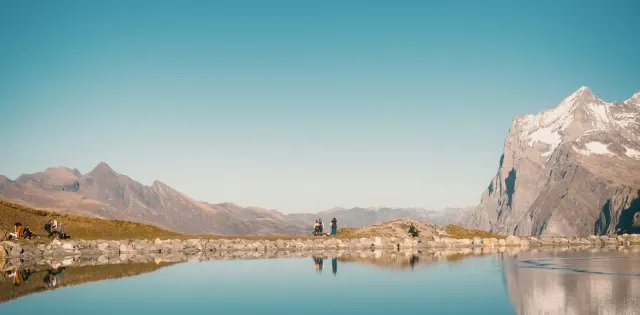 A still alpine lake reflecting a snowy peak and hikers on the shore